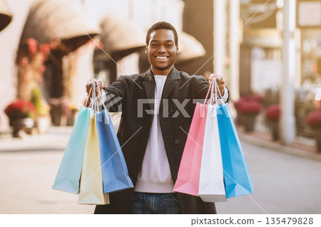 Beginning of sales season and Black Friday for shopaholic before holidays. Happy millennial african american man shows colored bags in camera near modern city mall after shopping, free space Beginning of sales season and Black Friday for shopaholic before holidays. Happy millennial african american man shows colored bags in camera near modern city mall after shopping, free space 135479828