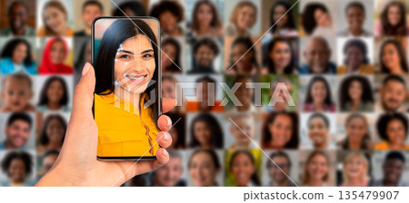 A person holds a smartphone displaying a facial recognition scan of their face. The background features a collage of diverse faces, showcasing the technology's application in identification. A person holds a smartphone displaying a facial recognition scan of their face. The background features a collage of diverse faces, showcasing the technology's application in identification. 135479907