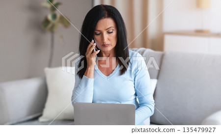 People And Technology Concept. Portrait of serious mature lady sitting on the couch at living room, talking on mobile phone and using laptop. Focused busy businesswoman working at home office 135479927