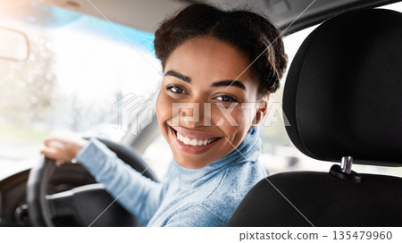 Driver turns to passenger seat and smiles at camera. Happy millennial african american woman taxi driver sitting in car holding to steering wheel, next to smartphone with blank screen, panorama Driver turns to passenger seat and smiles at camera. Happy millennial african american woman taxi driver sitting in car holding to steering wheel, next to smartphone with blank screen, panorama 135479960