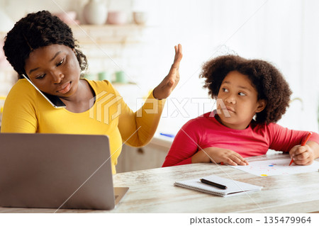 Busy Working African American Mother Ignoring Her Little Daughter Asking For Attention While Drawing In Kitchen. Young Woman Showing Not Now Gesture With Palm, Talking On Cellphone And Using Laptop 135479964