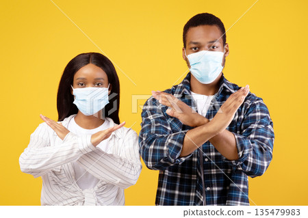 Stop virus epidemic and social distancing. Serious millennial african american guy and lady in protective masks cross arms, showing sign, isolated on yellow background, studio shot, copy space 135479983