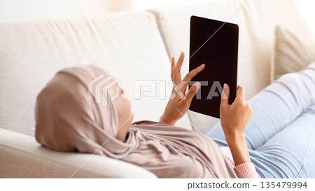 African American woman in hijab with tablet computer lying on sofa at home, mockup for design on screen. Muslim lady studying or working online, watching movie or chatting with friend 135479994