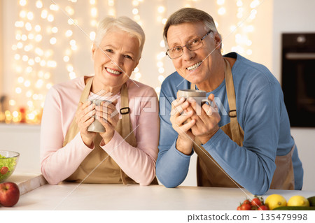 Happy Senior Couple Drinking Coffee Holding Mugs Celebrating Christmas Eve Together Sitting In Kitchen At Home, Smiling To Camera. Homey New Year Celebration. Elderly Spouses Enjoying Xmas Holiday 135479998