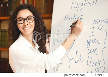 Lecture. Closeup portrait of cheerful smiling female tutor wearing glasses teaching English language, writing grammar rules on the board with marker, looking back at her students at classroom Lecture. Closeup portrait of cheerful smiling female tutor wearing glasses teaching English language, writing grammar rules on the board with marker, looking back at her students at classroom 135479999