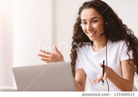 Excited young beautiful brunette woman having video interview with employer from home, using headset and laptop, looking at computer screen, talking and gesturing, closeup, copy space Excited young beautiful brunette woman having video interview with employer from home, using headset and laptop, looking at computer screen, talking and gesturing, closeup, copy space 135480015