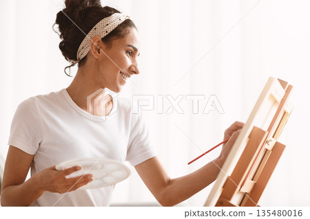 Young smiling woman artist painting at home, using easel, palette and paintbrush, side view, copy space. Closeup of creative lady drawing while having free time during quarantine Young smiling woman artist painting at home, using easel, palette and paintbrush, side view, copy space. Closeup of creative lady drawing while having free time during quarantine 135480016