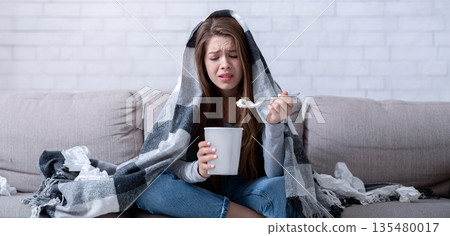Stress eating concept. Upset lady eating ice cream and crying under warm plaid at home. Lonely young woman watching sad romantic movie, going through breakup, having broken heart 135480017