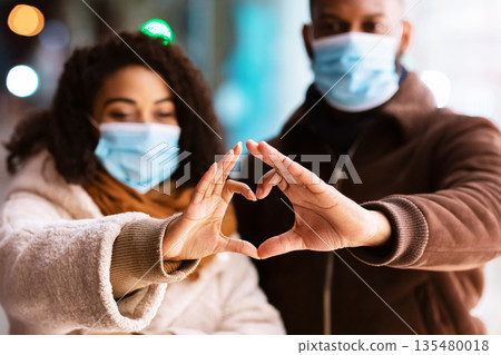 Love During Pandemic Concept. Portrait of african american couple wearing medical face masks making heart shape with hands walking together in the evening, selective focus. Connection And Support Love During Pandemic Concept. Portrait of african american couple wearing medical face masks making heart shape with hands walking together in the evening, selective focus. Connection And Support 135480018