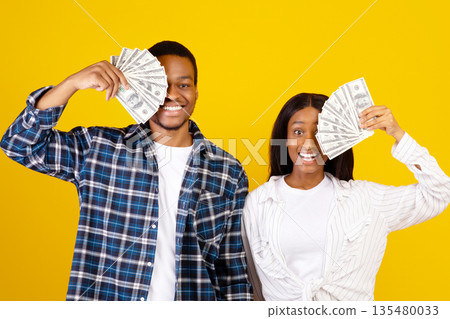 Online winning, cash back and income. Happy young african american lady and guy close their eyes with dollars and rejoice to victory at self-isolation, isolated on yellow background, studio shot Online winning, cash back and income. Happy young african american lady and guy close their eyes with dollars and rejoice to victory at self-isolation, isolated on yellow background, studio shot 135480033
