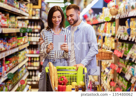 Young lovely couple with shopping cart full of healthy eco products looking into their grocery list at supermarket. Smiling man embaricng woman while buying products at mall together 135480062