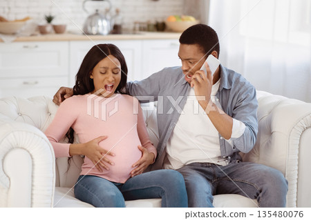 Expectant black woman having prenatal contractions, her boyfriend reaching doctor on smartphone from home. African American lady giving birth to baby, her husband calling for medical assistance 135480076
