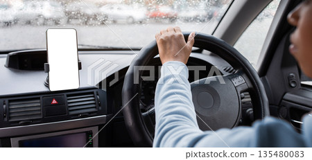 Modern gadgets for driving, working in city and traveling. Serious millennial beautiful african american woman holding steering wheel and using smartphone, driving car in rain, cropped, empty space Modern gadgets for driving, working in city and traveling. Serious millennial beautiful african american woman holding steering wheel and using smartphone, driving car in rain, cropped, empty space 135480083