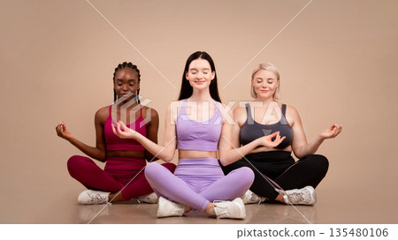 Beautiful body positive women of different size and appearances meditating together indoors, young happy multiethnic females in sportswear sitting in lotus pose with eyes closed over beige background 135480106