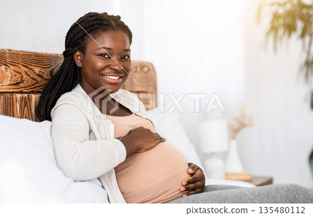 Enjoying Pregnancy. Cheerful African American Pregnant Woman Resting In Bed And Touching Her Belly, Happy Black Lady Relaxing In Cozy Bedroom At Home, Cuddling Tummy And Smiling At Camera, Copy Space Enjoying Pregnancy. Cheerful African American Pregnant Woman Resting In Bed And Touching Her Belly, Happy Black Lady Relaxing In Cozy Bedroom At Home, Cuddling Tummy And Smiling At Camera, Copy Space 135480112