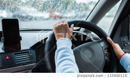Online map, navigator in mobile device and trip in city in bad weather. Young african american female drives car and holds steering wheel, view of windshield with raindrops, cropped, pov, empty space Online map, navigator in mobile device and trip in city in bad weather. Young african american female drives car and holds steering wheel, view of windshield with raindrops, cropped, pov, empty space 135480118