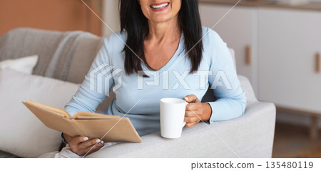 Leisure, Rest And Relax Concept. Portrait of smiling mature woman reading book and holding mug of coffee, sitting on the sofa in living room in her apartment, enjoying weekend and looking at camera Leisure, Rest And Relax Concept. Portrait of smiling mature woman reading book and holding mug of coffee, sitting on the sofa in living room in her apartment, enjoying weekend and looking at camera 135480119