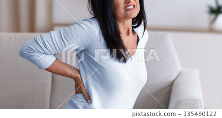 Portrait of sad mature woman having back pain while sitting on the couch at home, suffering from tension and acute spinal ache, feeling uncomfortable, stressed lady tired of work 135480122