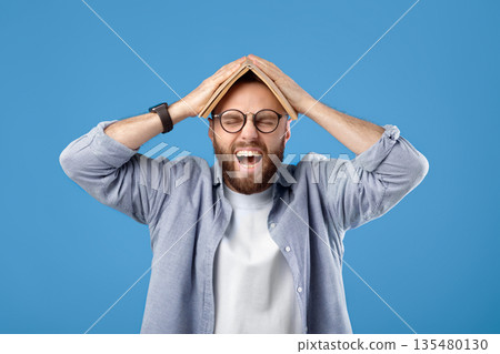 Furious young man putting book on his head and screaming on blue studio background. Bearded millennial guy tired of studying, cannot do difficult home assignment or meet deadline 135480130