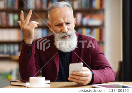 Angry senior man with grey hair and long beard using mobile phone while drinkin coffee at cafe, having negative face expression and gesturing, got bad news, having digital tablet by him, copy space 135480153