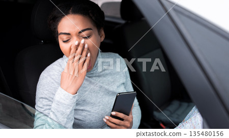 Road in early morning, create route and overwork in big traveling. Tired cute millennial african american lady driver yawns and holds smartphone sitting at steering wheel in own car, copy space Road in early morning, create route and overwork in big traveling. Tired cute millennial african american lady driver yawns and holds smartphone sitting at steering wheel in own car, copy space 135480156