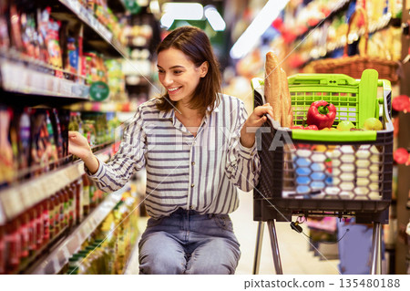 At The Supermarket. Portrait Of Smiling Millennial Lady With Shopping Cart In Wholesale Market Buying Groceries Food Nutrition, Taking Products From Shelves In Store, Low Angle View At The Supermarket. Portrait Of Smiling Millennial Lady With Shopping Cart In Wholesale Market Buying Groceries Food Nutrition, Taking Products From Shelves In Store, Low Angle View 135480188