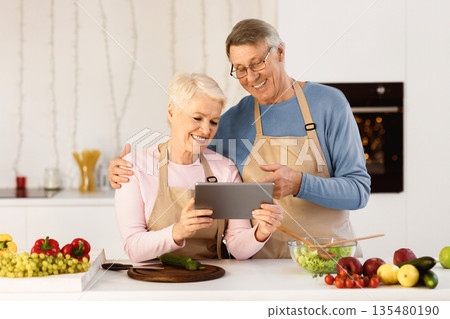 Cheerful Elderly Couple Using Digital Tablet Cooking Together, Searching Food Recipes Online Preparing Dinner In Modern Kitchen At Home. Cooking Application Concept. Cheerful Elderly Couple Using Digital Tablet Cooking Together, Searching Food Recipes Online Preparing Dinner In Modern Kitchen At Home. Cooking Application Concept. 135480190