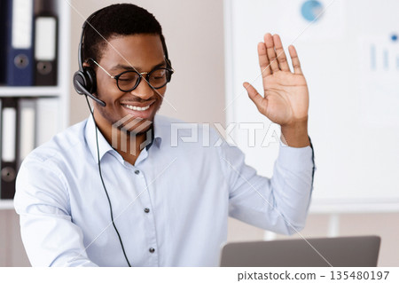 Office worker concept. Happy african american guy consultant with headset waving at laptop screen, office interior, empty space. Cheerful black man employer working online, wearing glasses Office worker concept. Happy african american guy consultant with headset waving at laptop screen, office interior, empty space. Cheerful black man employer working online, wearing glasses 135480197