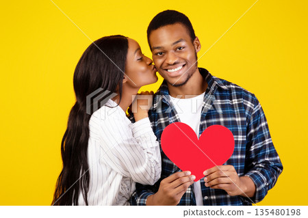 Couple in love, happy emotions and valentine day. Millennial african american woman kissing smiling man, holding red heart and celebrate anniversary, isolated on yellow background, studio shot Couple in love, happy emotions and valentine day. Millennial african american woman kissing smiling man, holding red heart and celebrate anniversary, isolated on yellow background, studio shot 135480198