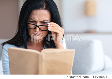Portrait of confused senior woman squinting to see more clearly, wearing and touching eyeglasses, trying to read paper book, having difficulties seeing text because of bad vision problems and issues Portrait of confused senior woman squinting to see more clearly, wearing and touching eyeglasses, trying to read paper book, having difficulties seeing text because of bad vision problems and issues 135480219