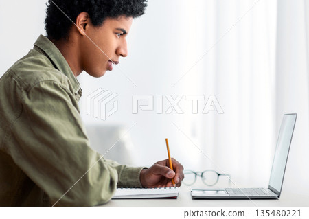 Side view of millennial black student sitting at desk with laptop, making notes in copybook, learning remotely from home, free space. African American adolescent on homeschooling 135480221