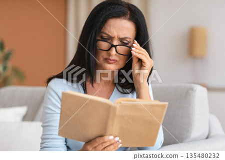 Portrait of mature woman touching glasses trying to read book, having difficulties seeing text because of vision problems. Negative emotion facial expression feelings reaction, health issues Portrait of mature woman touching glasses trying to read book, having difficulties seeing text because of vision problems. Negative emotion facial expression feelings reaction, health issues 135480232