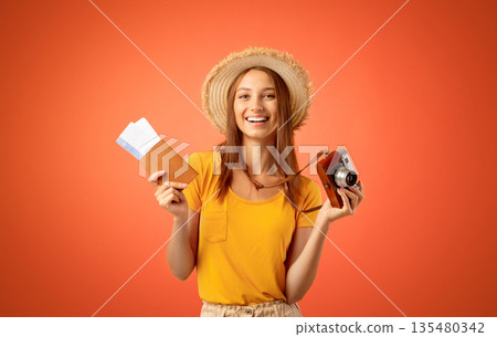 Smiling young woman in summer hat holding camera and passport with flight tickets in, yellow studio background Smiling young woman in summer hat holding camera and passport with flight tickets in, yellow studio background 135480342