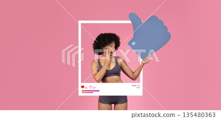 A young woman in a cropped top and shorts expresses surprise while pointing at a large thumbs-up symbol. The vibrant pink backdrop enhances her excitement, showcasing social media interaction. 135480363