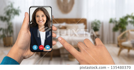 A person is holding a smartphone displaying a smiling face during an online conversation at home. The cozy room features a bed and potted plants, emphasizing a relaxed atmosphere. A person is holding a smartphone displaying a smiling face during an online conversation at home. The cozy room features a bed and potted plants, emphasizing a relaxed atmosphere. 135480370