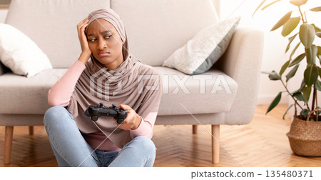 Full length portrait of upset black woman in hijab losing computer game, sitting with joystick on floor at home. Unhappy Muslim lady spending time playing online videogames Full length portrait of upset black woman in hijab losing computer game, sitting with joystick on floor at home. Unhappy Muslim lady spending time playing online videogames 135480371