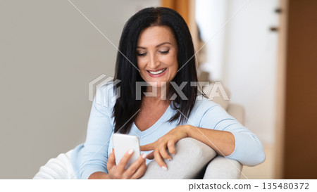 Leisure At Home. Smiling mature brunette woman using her cellphone, browsing internet, checking social media, sitting on the sofa in living room. Happy lady texting her friends, watching video online Leisure At Home. Smiling mature brunette woman using her cellphone, browsing internet, checking social media, sitting on the sofa in living room. Happy lady texting her friends, watching video online 135480372