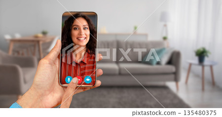A person participates in a video call using a smartphone. The setting is a cozy home with a comfortable sofa in the background and natural light coming in. A person participates in a video call using a smartphone. The setting is a cozy home with a comfortable sofa in the background and natural light coming in. 135480375