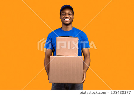 Best Service Concept. Smiling black delivery man in blue cap and unform holding pile of cardboard boxes standing isolated over orange studio background. African american courier carrying packages Best Service Concept. Smiling black delivery man in blue cap and unform holding pile of cardboard boxes standing isolated over orange studio background. African american courier carrying packages 135480377