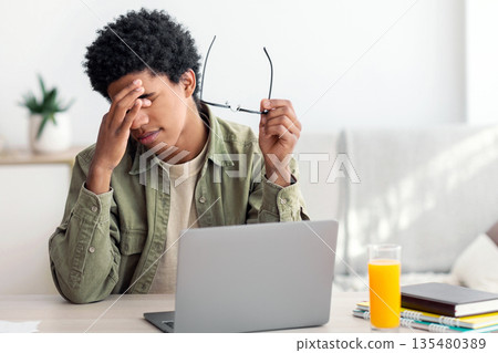 Black teenager feeling tired, rubbing irritated eyes, sitting at desk with laptop, exhausted from studying online. African American guy feeling fatigue from looking at screen, doing assignments on web Black teenager feeling tired, rubbing irritated eyes, sitting at desk with laptop, exhausted from studying online. African American guy feeling fatigue from looking at screen, doing assignments on web 135480389