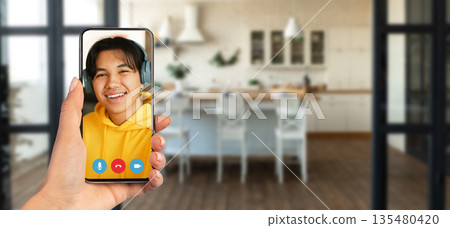 A person is holding a smartphone displaying a smiling individual on a video call. The setting is a cozy home with a modern kitchen visible in the background. 135480420