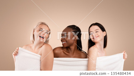 Beauty And Spa. Three Body Positive Multi-Ethnic Women With Different Size And Appearances Hiding Behind Bath Towel, Having Fun Together Over Beige Background, Enjoying Themselves, Copy Space 135480434