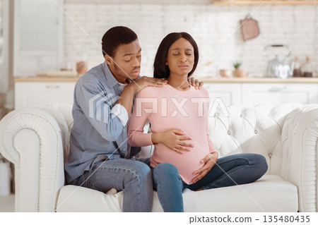 Worried black husband supporting pregnant wife with prenatal contractions at home. Beautiful African American woman suffering from pain, giving birth to baby with help of her partner Worried black husband supporting pregnant wife with prenatal contractions at home. Beautiful African American woman suffering from pain, giving birth to baby with help of her partner 135480435