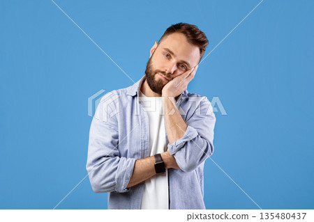 Bored young guy leaning on his hand and looking at camera over blue studio background. Tired millennial man feeling exhausted or overworked, not interested in something 135480437