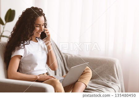 Happy young woman freelancer talking on mobile phone and using laptop, working from home during isolation, copy space. Curly lady having conversation on phone and typing on computer keyboard Happy young woman freelancer talking on mobile phone and using laptop, working from home during isolation, copy space. Curly lady having conversation on phone and typing on computer keyboard 135480450