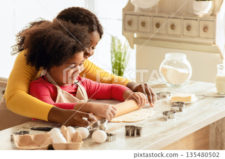 Loving Black Mom Teaching Daughter How To Roll Dough, Having Fun Together In Kitchen At Home, African American Lady Helping To Her Little Child While They Making Homemade Pastry, Free Space 135480502