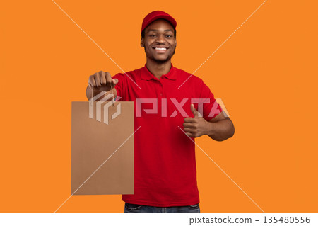 Portrait of african american delivery man in red uniform holding craft paper bag, showing thumbs up gesture, isolated on orange background. Design, mockup, blank space on package for advert Portrait of african american delivery man in red uniform holding craft paper bag, showing thumbs up gesture, isolated on orange background. Design, mockup, blank space on package for advert 135480556