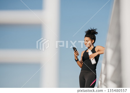 Social media and sport. Smiling athletic african american young woman in fashion sportswear holding bottle of water and looking at smartphone at stadium, free space Social media and sport. Smiling athletic african american young woman in fashion sportswear holding bottle of water and looking at smartphone at stadium, free space 135480557