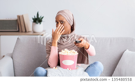 Young black woman in hijab watching scary movie, covering her mouth in horror, eating popcorn on sofa at home. Millennial Muslim lady feeling afraid, switching thriller off with remote control Young black woman in hijab watching scary movie, covering her mouth in horror, eating popcorn on sofa at home. Millennial Muslim lady feeling afraid, switching thriller off with remote control 135480558