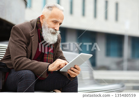 Stylish grey-haired elderly man sitting on the bench by office building, using digital tablet, checking his agenda, copy space. Aged man using pad on the street, websurfing while resting Stylish grey-haired elderly man sitting on the bench by office building, using digital tablet, checking his agenda, copy space. Aged man using pad on the street, websurfing while resting 135480559